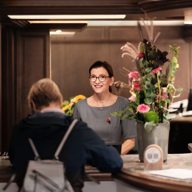 Die Rezeption im Traube Tonbach: Eine Mitarbeiterin unterhält sich mit einem Gast. Neben ihr steht ein Blumenstrauß in einer Vase.