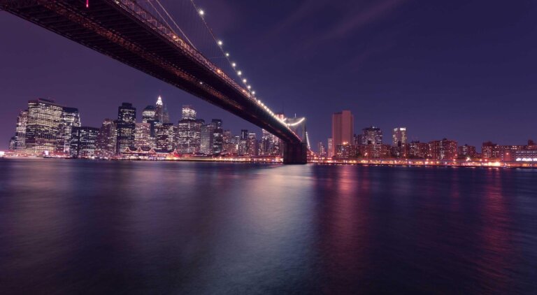 Brooklyn Bridge bei Nacht mit Skyline von New York City – spektakuläre Lichter der Metropole spiegeln sich im East River.