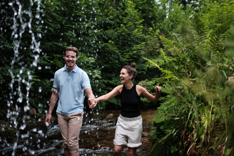 Ein lachendes Paar läuft Hand in Hand durch einen Bach unter einem Wasserfall.