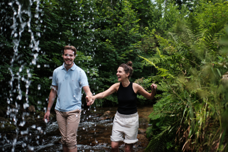 Ein lachendes Paar läuft Hand in Hand durch einen Bach unter einem Wasserfall.