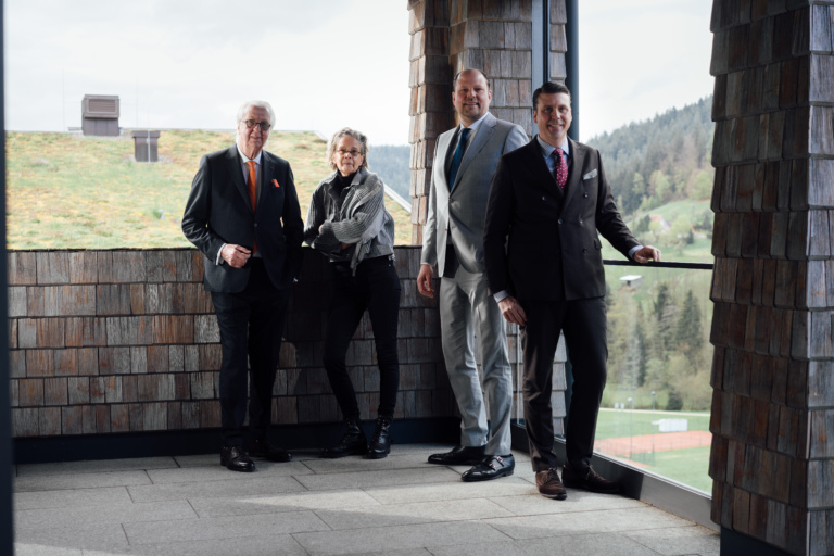 Vier Personen in eleganter Kleidung posieren auf einer Terrasse des Hotel Traube Tonbach mit Blick auf die umliegende Natur und Wälder im Schwarzwald.
