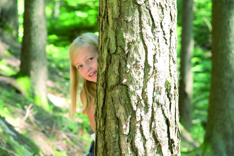 Ein blondes Mädchen schaut lachend hinter einem Baum im Wald im Schwarzwald hervor.