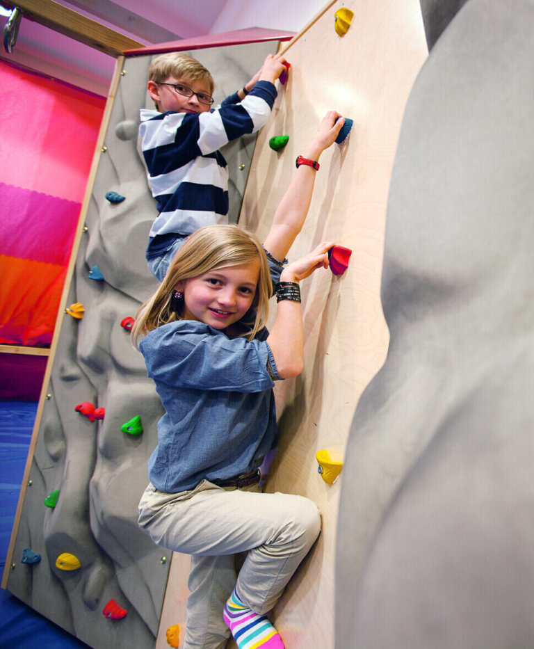 Kinder klettern an einer Indoor-Kletterwand im Hotel Traube Tonbach und lächeln sich.