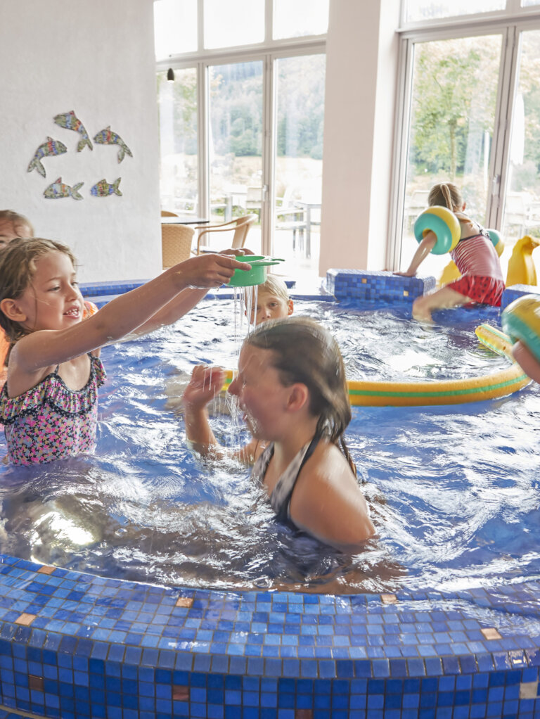 Mehrere Kinder spielen im Innenpool mit blauen Fliesen und Wasserspielzeug.