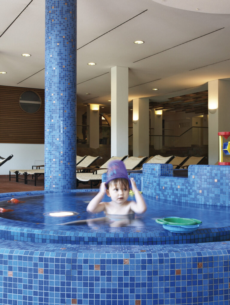 Ein kleines Kind spielt im blauen Kinderbecken des Hotel Traube Tonbach mit Eimer und Spielzeug, im Hintergrund Liegestühle im Wellnessbereich.