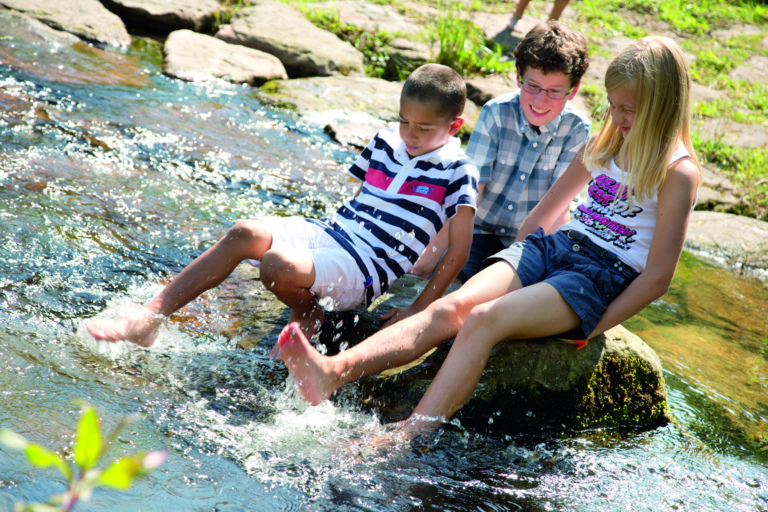 Drei Kinder sitzen lachend am Bach und spielen mit den Füßen im Wasser.