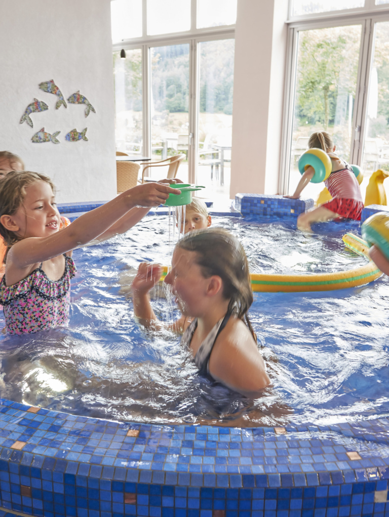 Mehrere Kinder spielen im Innenpool mit blauen Fliesen und Wasserspielzeug.