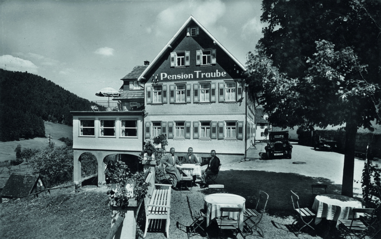 Schwarz-weißes Bild eines Gasthauses mit der Schild "Pension Traube". Sechs Personen sitzen an einem Tisch im Freien, umgeben von Stühlen und Tischen.