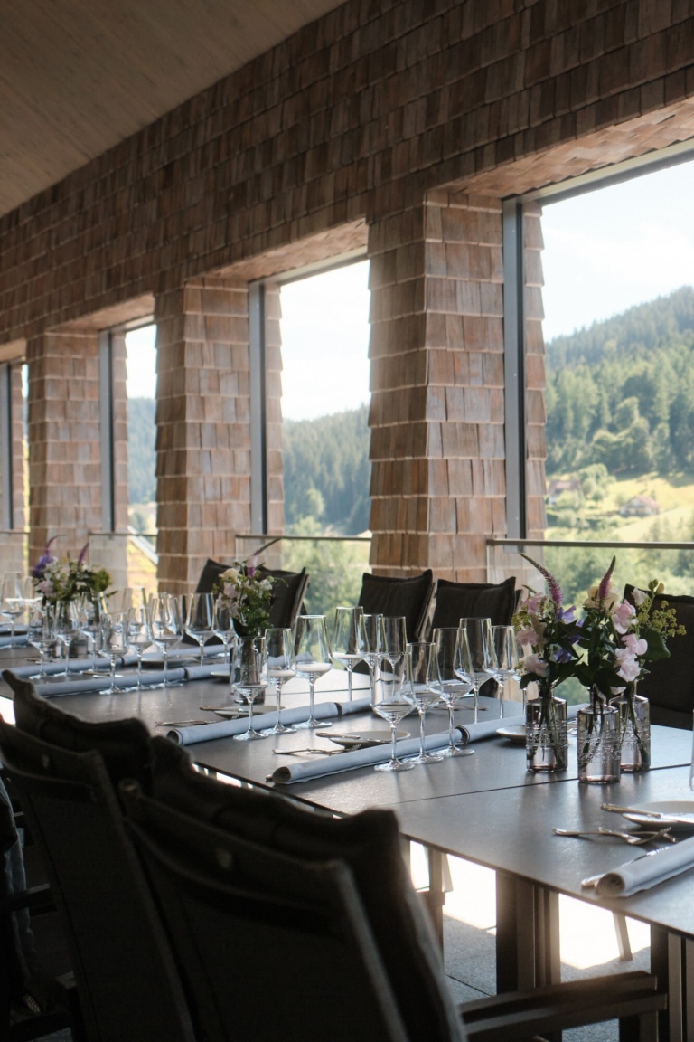 Ein gedeckter Tisch mit Gläsern und Blumen in einer modernen Restaurantumgebung mit großen Fenstern, die auf eine bewaldete Landschaft im Schwarzwald blicken.