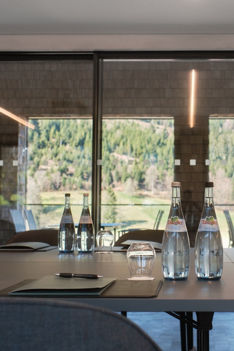 Konferenzraum mit einem Tisch, auf dem mehrere Glasflaschen mit Wasser stehen. Im Hintergrund sind grüne Bäume und ein Fenster sichtbar.