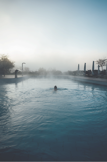 Eine Person schwimmt in einem dampfenden, blauen Pool in eniem Luxushotel im Schwarzwald. Im Hintergrund sind Bäume und Säulen sichtbar, umgeben von Nebel.