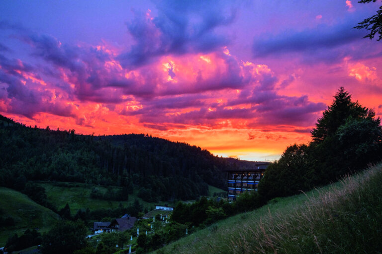 Landschaft im Schwarzwald mit bewaldeten Hügeln und einem dramatischen Sonnenuntergang, der den Himmel in lila und orange Tönen färbt.
