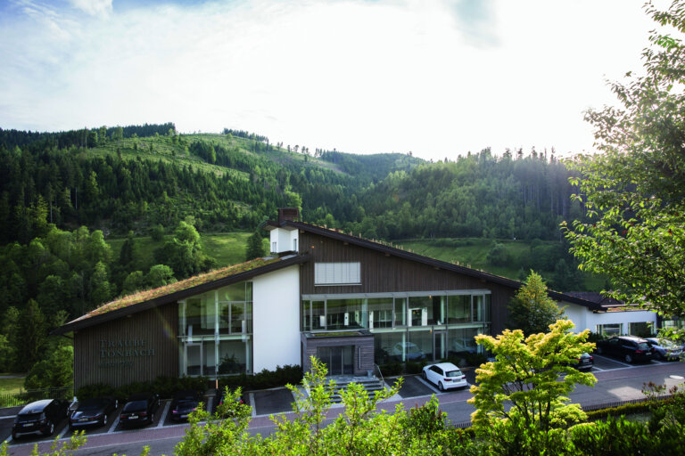 Ein modernes Gebäude mit Holzverkleidung und großen Fenstern, umgeben von grünen Hügeln und Bäumen im Schwarzwald. Vor dem Gebäude gibt es Parkplätze mit Autos.