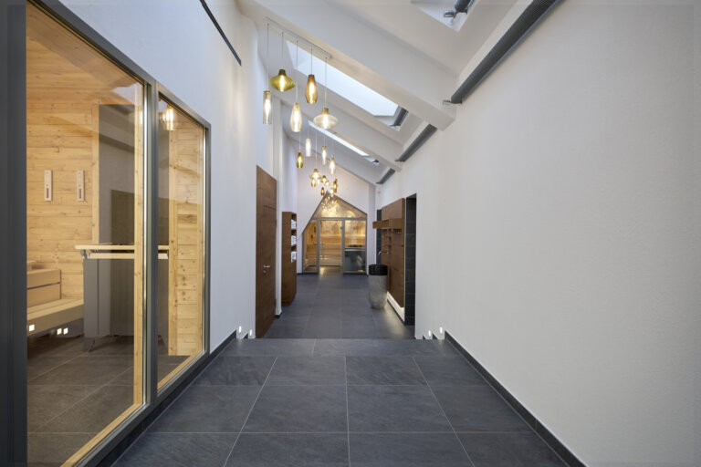 Blick auf einen langen Flur mit Holz- und Glastüren, hängenden Lampen und einem modernen, minimalistischen Design, links ist ein Saunabereich mit Holzeinrichtung.