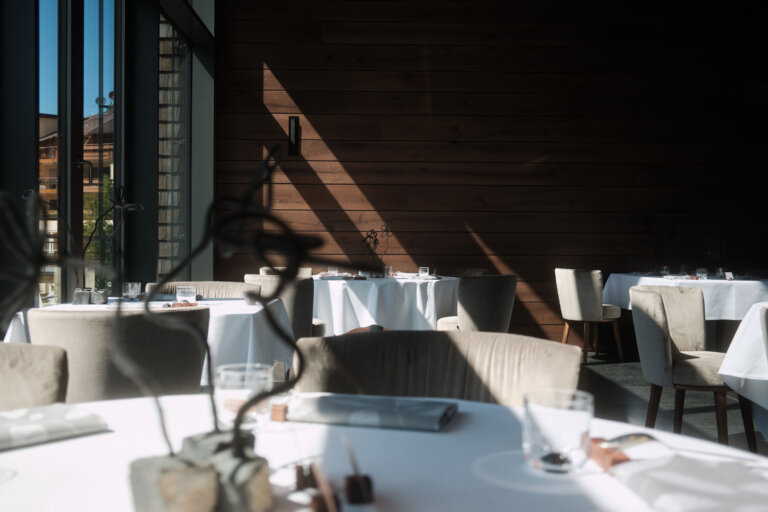 Ein eleganter Restaurantbereich eines Luxushotels mit runden Tischen, die mit weißen Tischdecken gedeckt sind. Stühle mit hellbeigem Stoff sind um die Tische gruppiert.