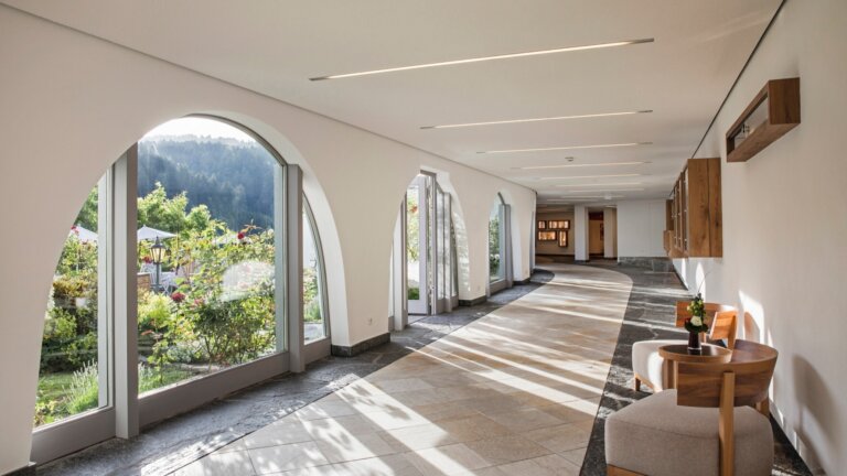 Ein heller Hotelflur mit großen Rundbogenfenstern mit Ausblick ins Grüne und einer gemütlichen Sitzecke.