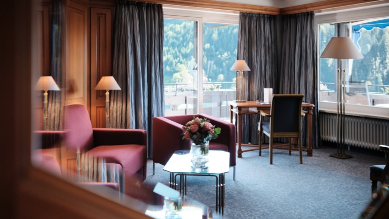 Ein Hotelzimmer mit einem Glastisch, Blumenstrauß, Schreibtisch vor Fensterfront und einer gemütlichen Sitzecke in Dunkelrot.