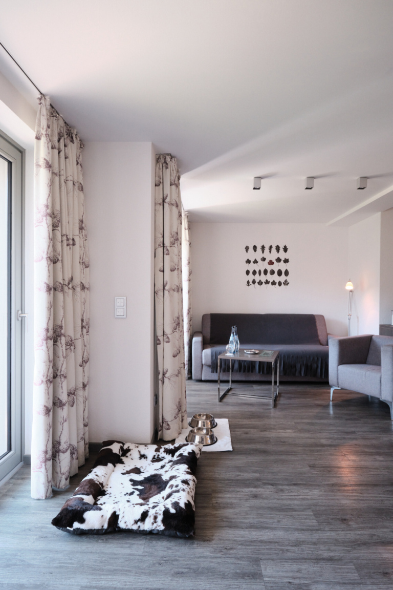 Ein hundefreundliches Hotelzimmer in einem 5-Sterne-Hotel im Schwarzwald mit einem grauen Sofa, einem Sessel, Holzboden und einem Hundebett.