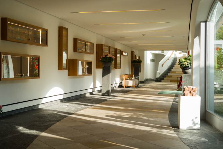 Ein heller Hotelflur mit Holzregalen an der Wand, Pflanzen in Töpfen und einer Treppe im Hintergrund. Der Boden ist aus Steinplatten.