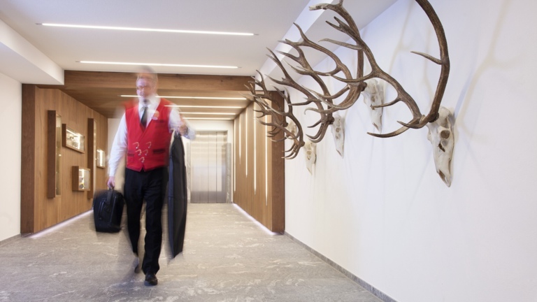 Eine Hotelflur mit Geweihen an der Wand, ein Mitarbeiter trägt die Hotel Uniform und hat was in seiner Hand. Im Hintergrund ist ein Aufzug zu sehen.