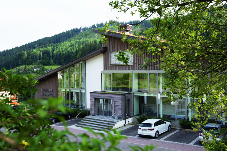 Ein modernes Gebäude mit großen Glasfenstern und Holzverkleidung, umgeben von Bäumen und einer grünen Landschaft. Davor gibt es einen parkplatz mit zwei Autos.