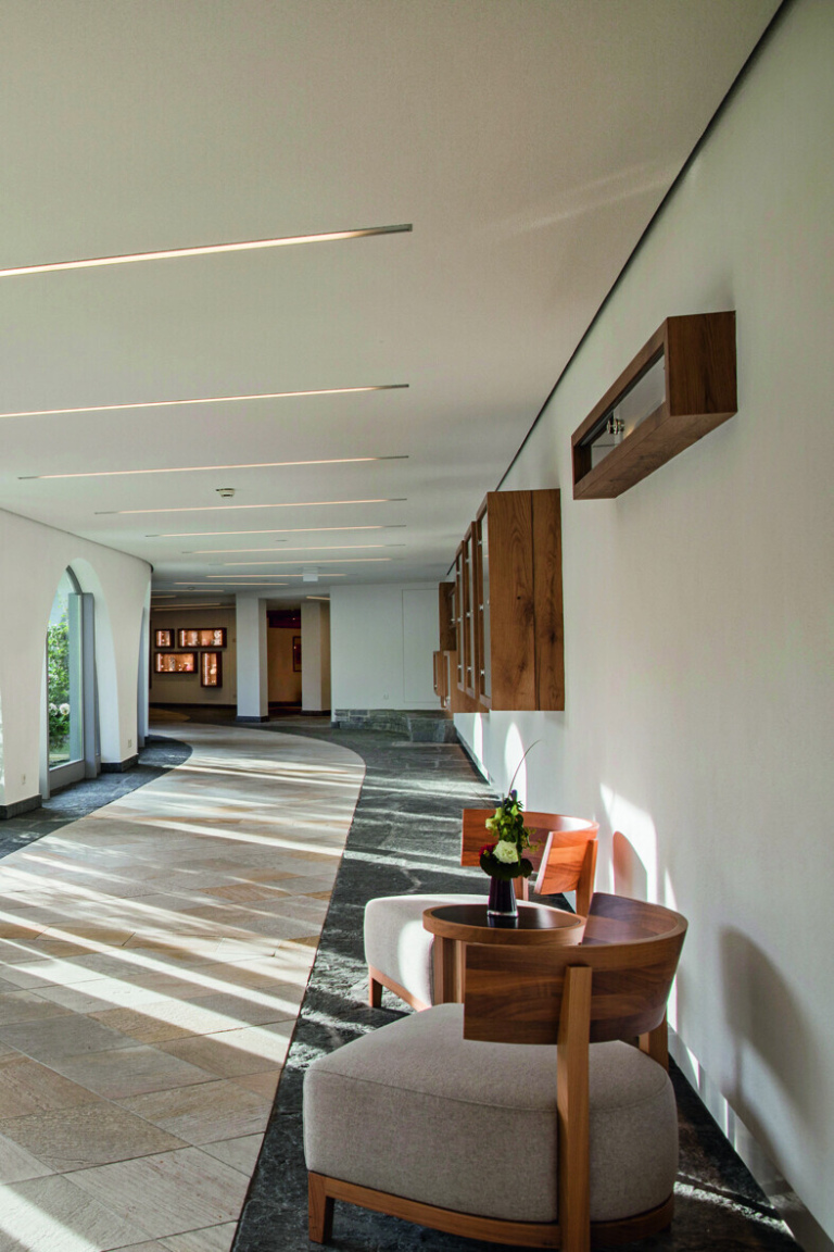 Helle Hotellobby im Schwarzwald mit einem geschwungenen Flur, Holzborden an der Wand, zwei modernen Sesseln und einem kleinen Tisch, auf dem eine Blumenvase steht.