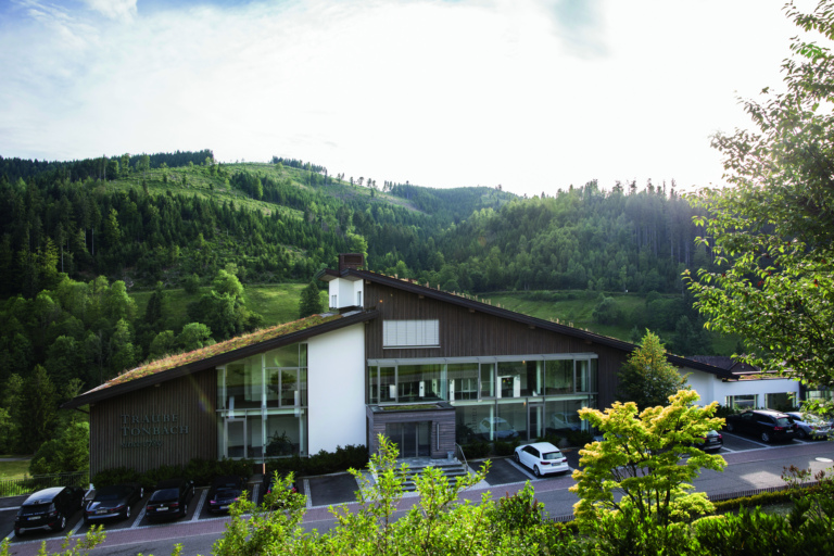 Ein modernes Gebäude mit Holzverkleidung und großen Fenstern, umgeben von grünen Hügeln und Bäumen im Schwarzwald. Vor dem Gebäude gibt es Parkplätze mit Autos.