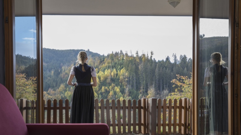 Eine blonde Frau steht auf einem Balkon im Hotelzimmer mit Blick auf bewaldete Hügel im Schwarzwald.