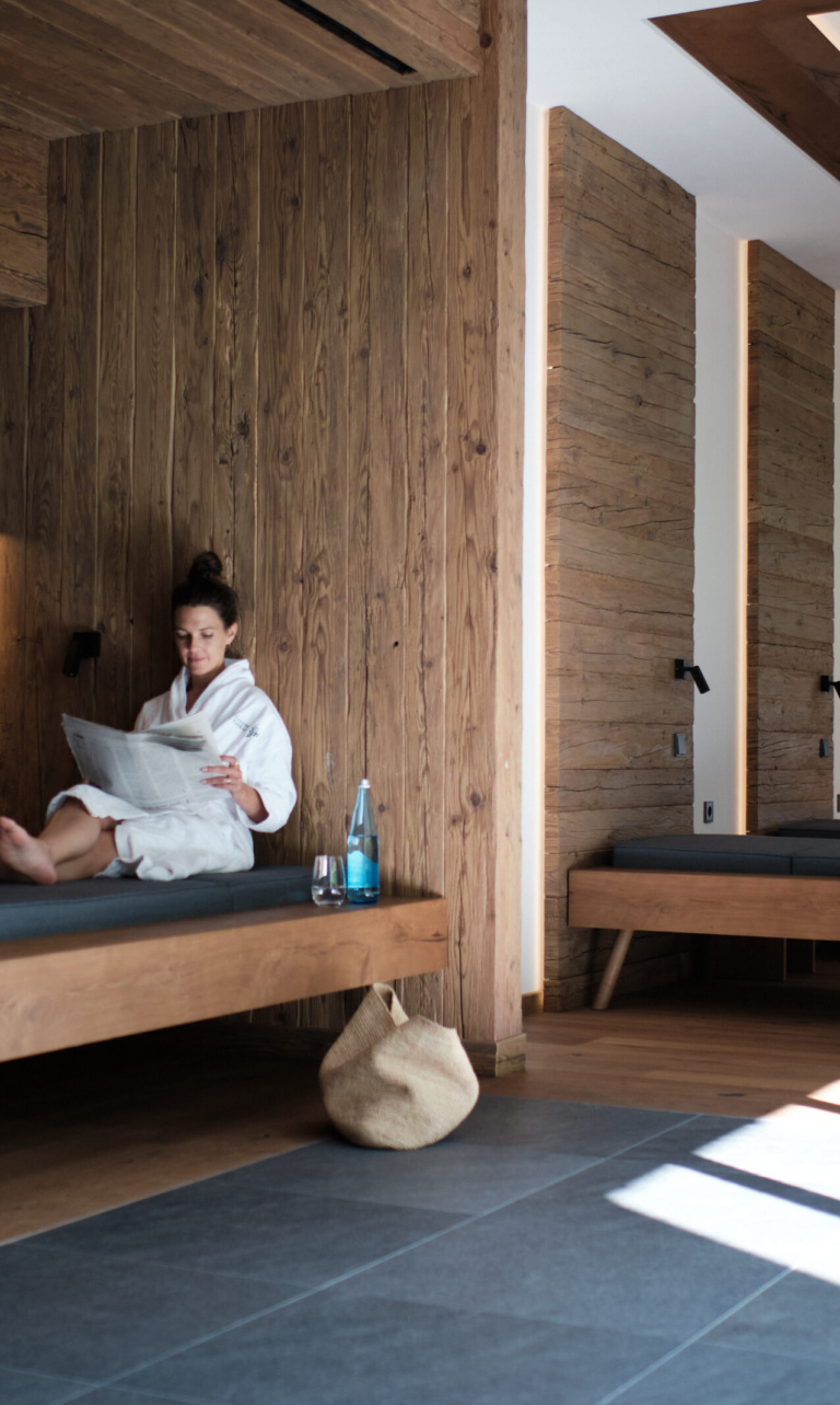 Eine Frau im Bademantel sitzt auf einer Liege in einem Spa-Bereich und liest eine Zeitung.