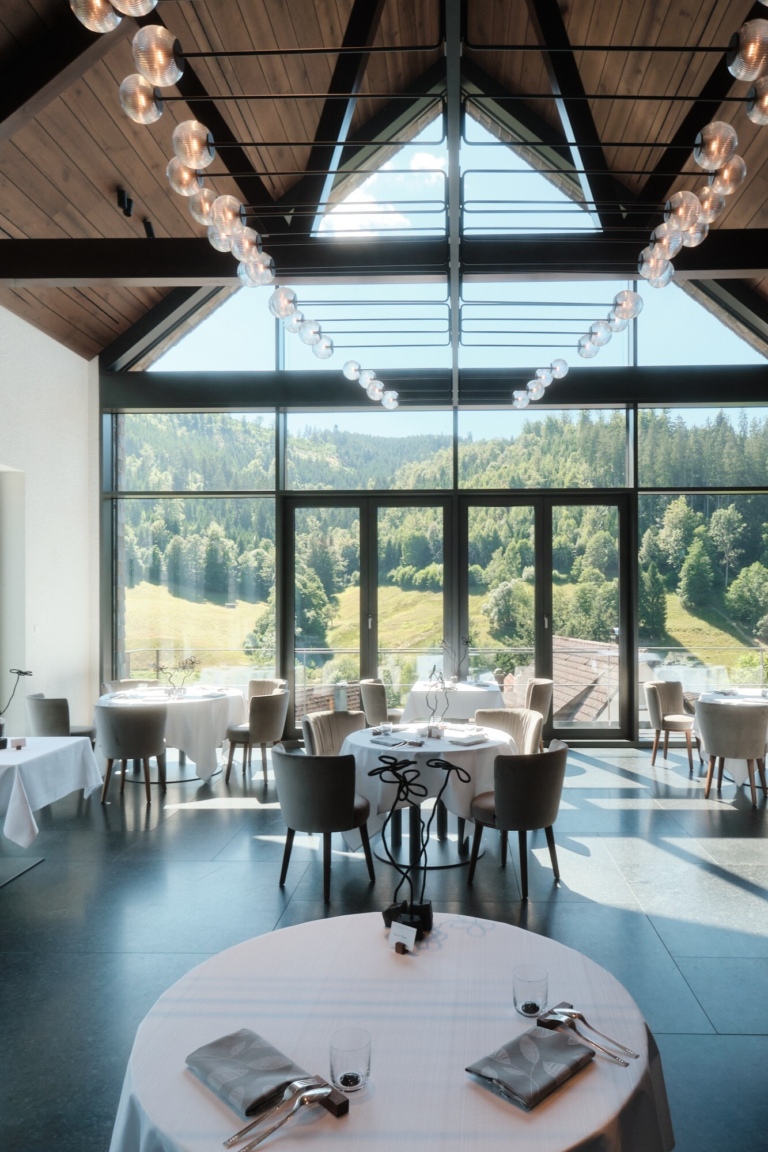 Ein Gourmet Restaurant mit Glasfront, runden Tischen und Blick auf bewaldete Hügel im Schwarzwald.