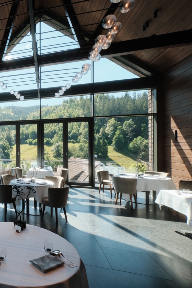 Ein Restaurant mit Glasfronten und Ausblick auf bewaldete Hügel im Schwarzwald. Runde Tische sind mit weißen Tischdecken gedeckt.