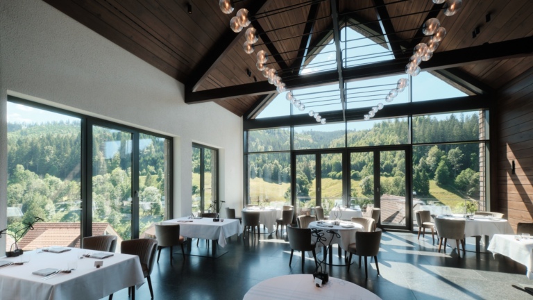 Ein großes Gourmet Restaurant mit großen Glasfronten, runden gedeckten Tischen mit weißen Tischdecken und Blick ins Grüne.