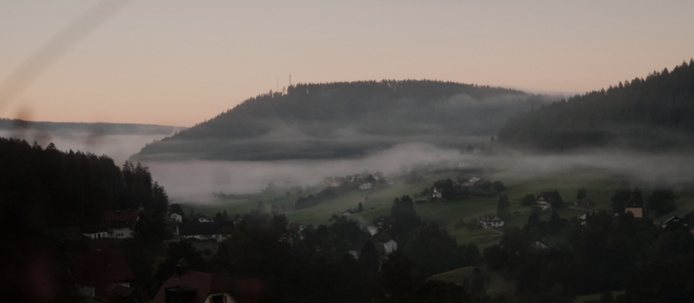 Blick auf den Schwarzwald bei Sonnenaufgang, mit Nebel über den Hügeln und verstreuten Häusern im Tal.