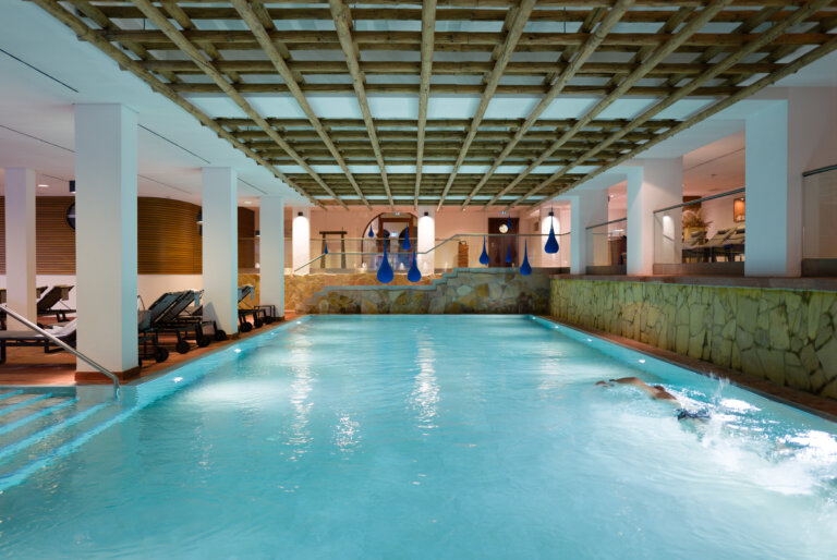 Ein sehr großer Innenpool im Hotel Traube Tonbach mit einer klaren Wasserfläche, Holzdecke und Liegestühlen, Gäste sind beim Schwimmen.
