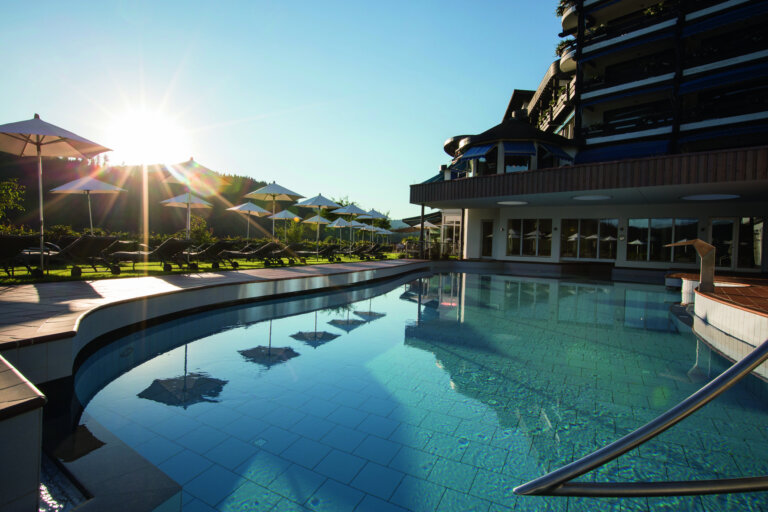 Ein beheizter Außenpool mit klarem Wasser, umgeben von Liegestühlen und Sonnenschirmen, durch die die Sonne scheint, im Hintergrund ist ein modernes Hotelgebäude mit mehreren Etagen.