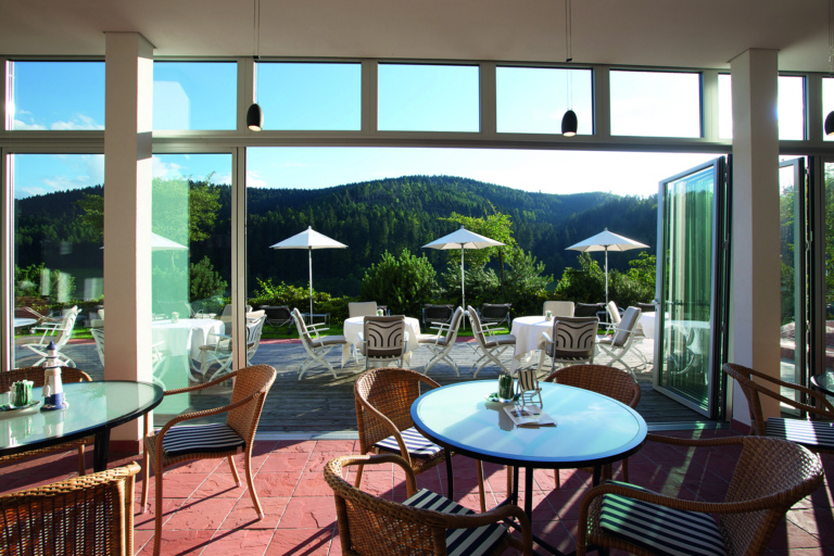 Eine helle Lounge mit Glasfront im Hotel Traube Tonbach, Blick auf Terrasse mit Sonnenschirmen und umliegende Wälder.