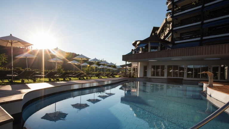 Ein beheizter Außenpool eines Hotels im Schwarzwald mit Sonnenschirmen und Liegen, die Sonne scheint.