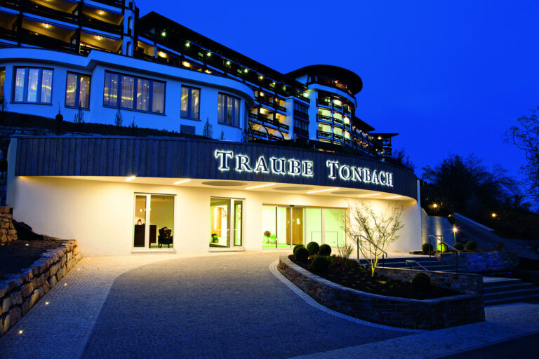 Das Gebäude von Traube Tonbach am späten Abend mit einem modernen, mehrstöckigen Design. Der Schriftzug '"TRAUBE TONBACH" ist prominent an der Fassade angebracht.