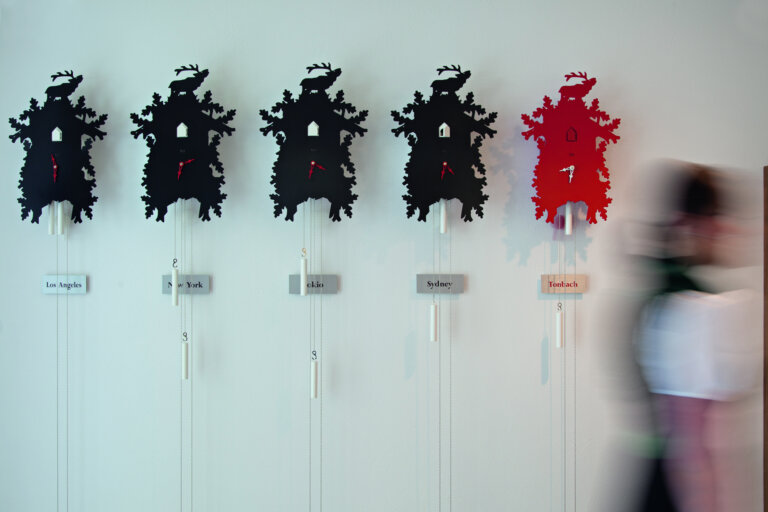 Moderne schwarze und rote Kuckucksuhren an einer weißen Wand, jede mit Städtenamen beschriftet.
