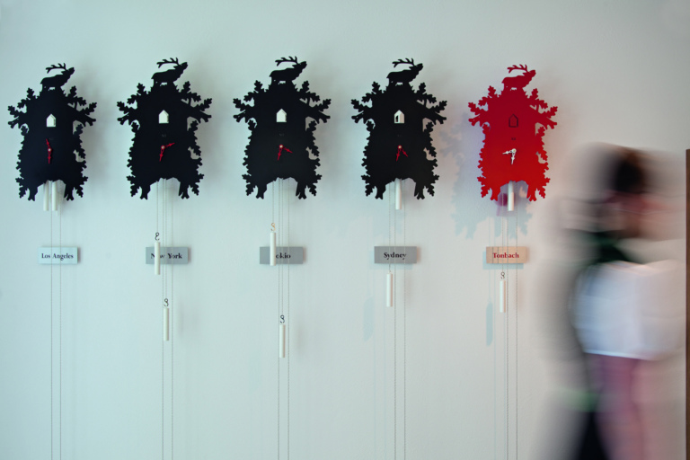 Moderne schwarze und rote Kuckucksuhren an einer weißen Wand, jede mit Städtenamen beschriftet.