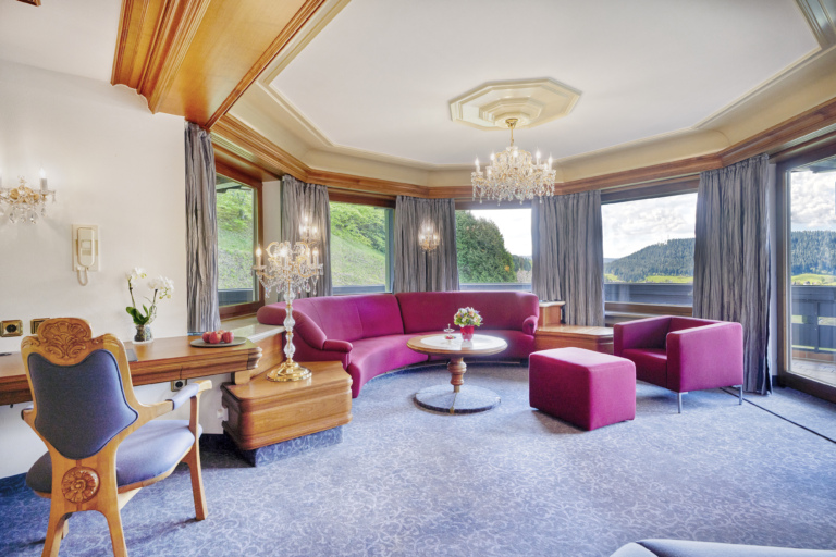 Ein elegantes Wohnzimmer in einer Hotel Suite mit Kronleuchter, Panoramafenstern und roten Sofas, stilvolles Interieur.