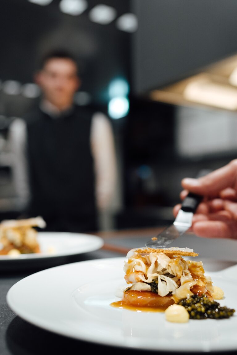 Ein Gourmetgericht auf einem weißen Teller, im Hintergrund steht ein Kellner in der Küche des Gourmet Restaurants in Traube Tonbach.