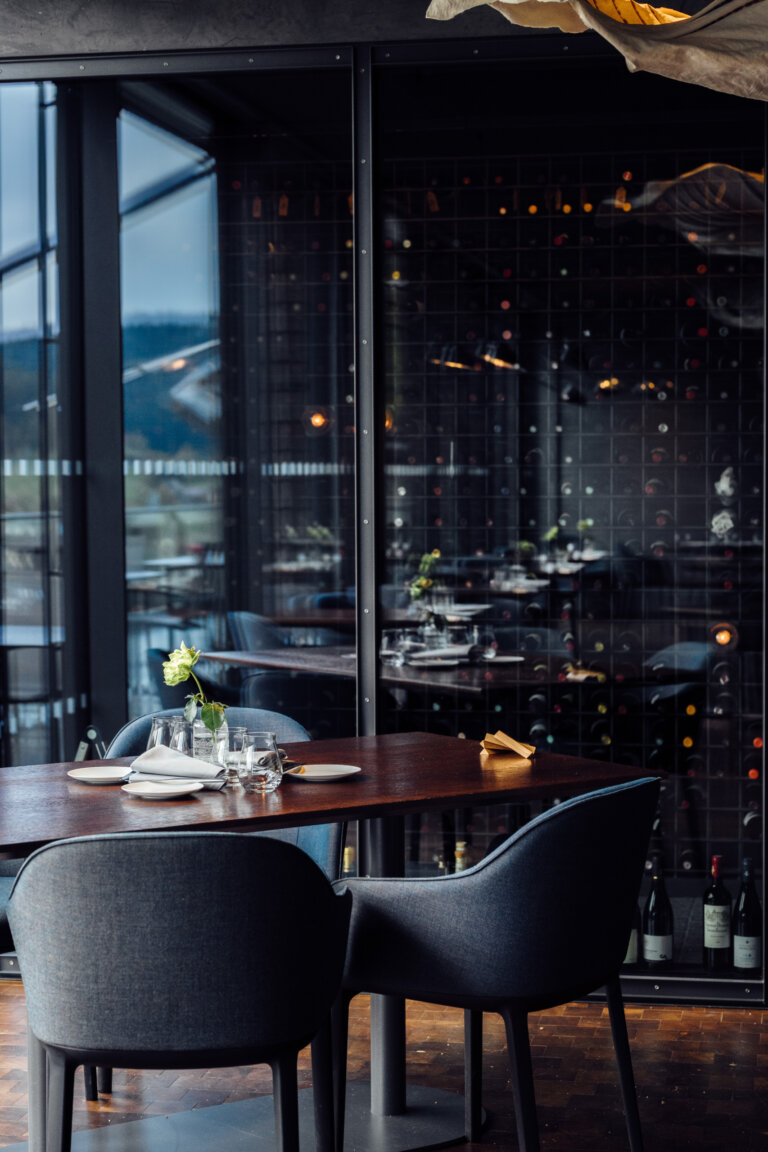 Ein moderner Restauranttisch mit zwei Stühlen vor einer Glaswand mit Weinflaschen.