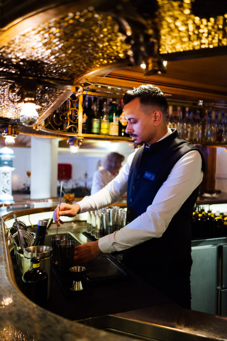 Ein Barkeeper in einem eleganten Hemd und Weste steht hinter einer Bar und bereitet Getränke vor. Im Hintergrund sind Regale mit Flaschen sichtbar.