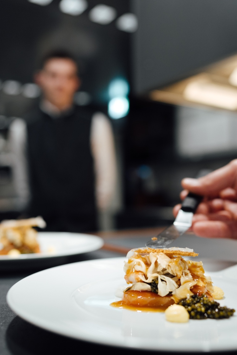 Ein Gourmetgericht auf einem weißen Teller, im Hintergrund steht ein Kellner in der Küche des Gourmet Restaurants in Traube Tonbach.