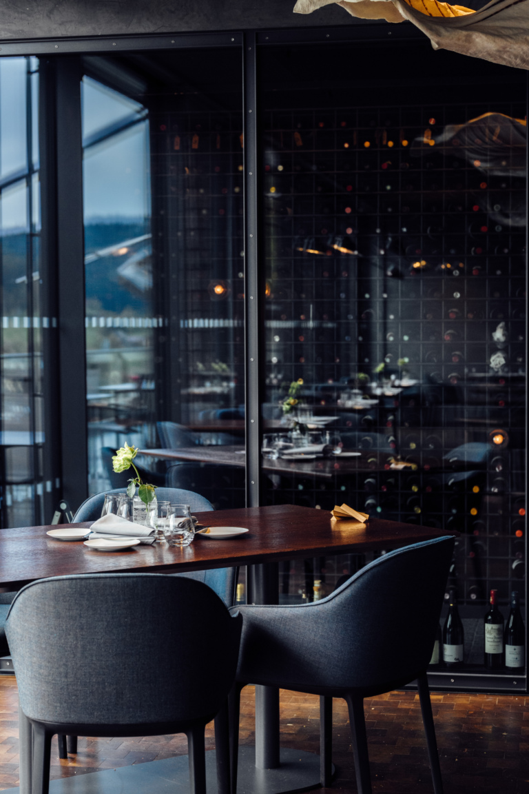 Ein eleganter Restaurantbereich mit einem Tisch, der mit Geschirr und Gläsern gedeckt ist. Zwei graue Stühle stehen um den Tisch. Im Hintergrund sind Weinfässer und eine große Fensterfront zu sehen.