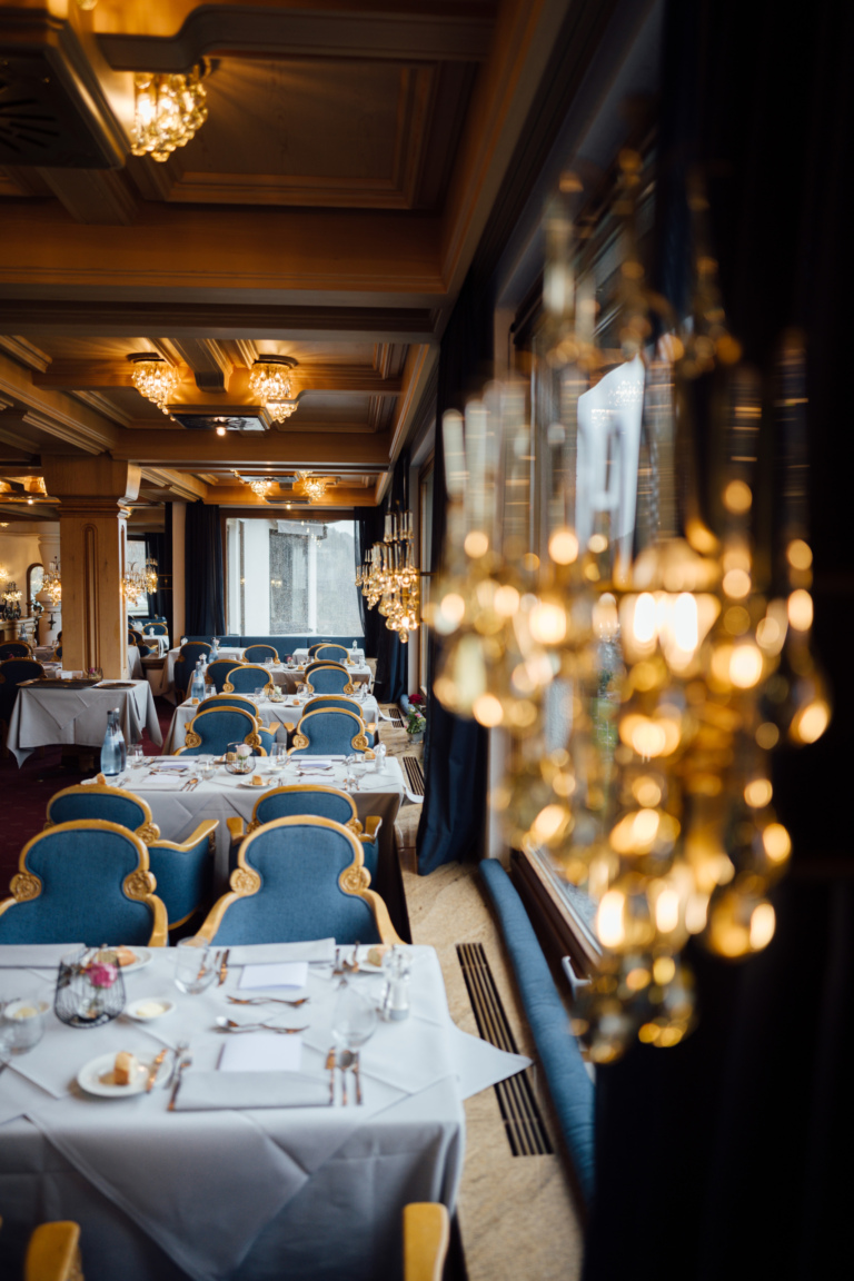 Ein elegantes Restaurant mit blauen Stühlen und weißen Tischdecken. Im Hintergrund sind Kristalllüster sichtbar, die sanft leuchten.