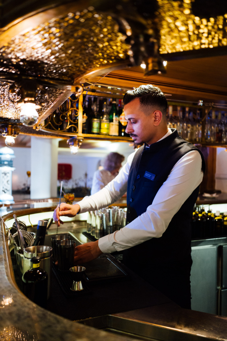 Ein Barkeeper in einem eleganten Hemd und Weste steht hinter einer Bar und bereitet Getränke vor. Im Hintergrund sind Regale mit Flaschen sichtbar.