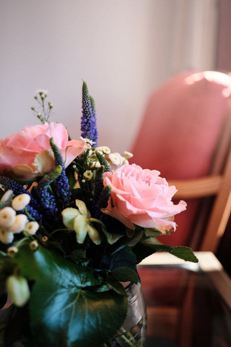 Ein Blumenstrauß mit rosa Rosen, blauen Blumen und grünen Blättern in einer Glasvase. Im Hintergrund ist ein korallenroter Stuhl sichtbar.
