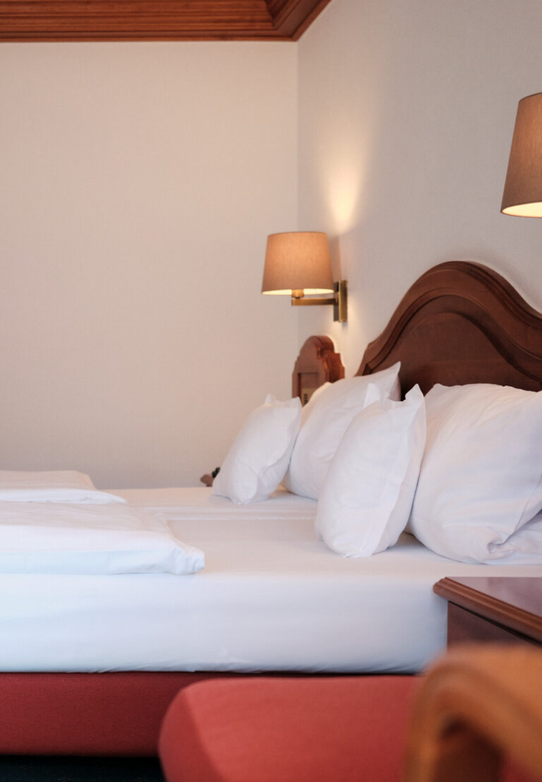 Ein Hotelzimmer mit einem Doppelbett, das mit weißen Bettwäschen bezogen ist. An der Wand hängen zwei Lampen und am Bett steht ein Nachttisch aus Holz.
